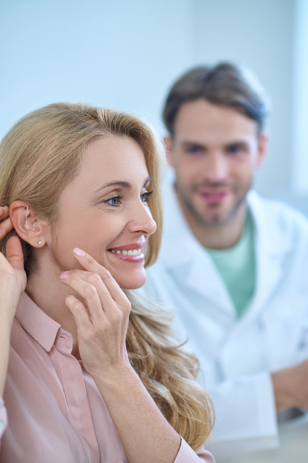 Femme souriante montrant son appareil auditif avec un audioprothesiste a l'arriere